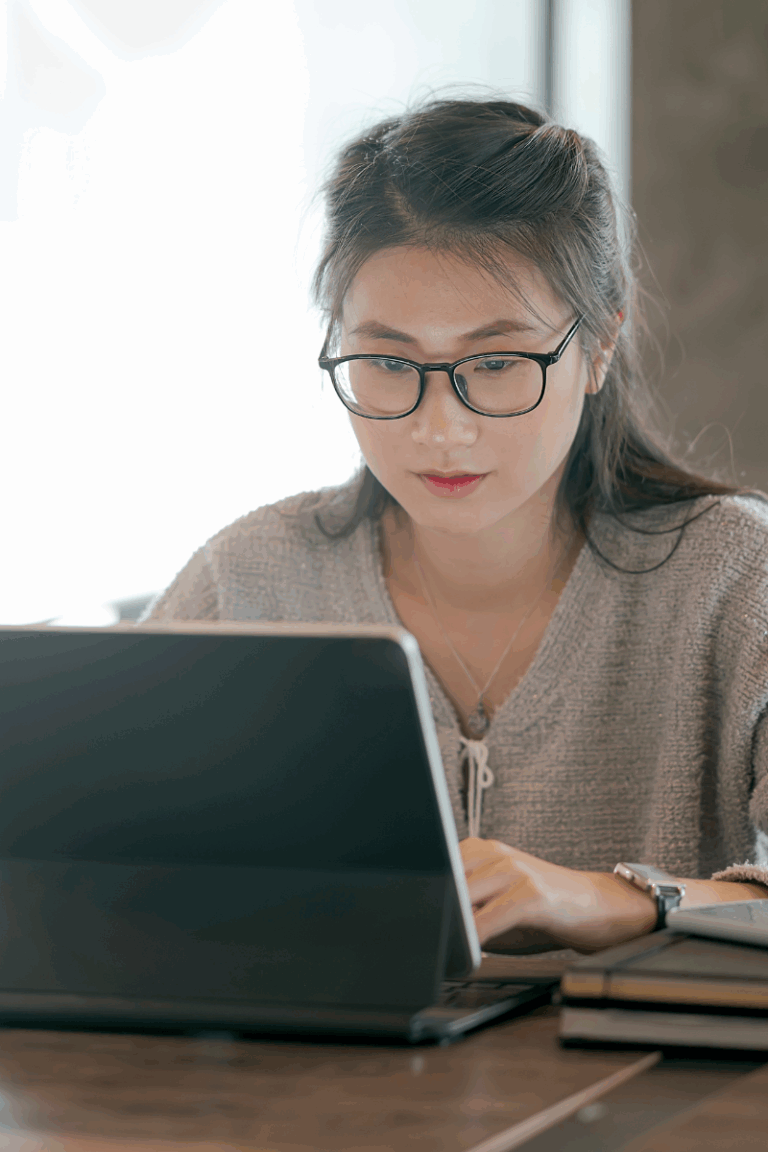 Woman using a tablet at a wooden desk with notebooks and soft lighting, illustrating the use of Aesthetic Notion Covers in a calm, organized workspace.