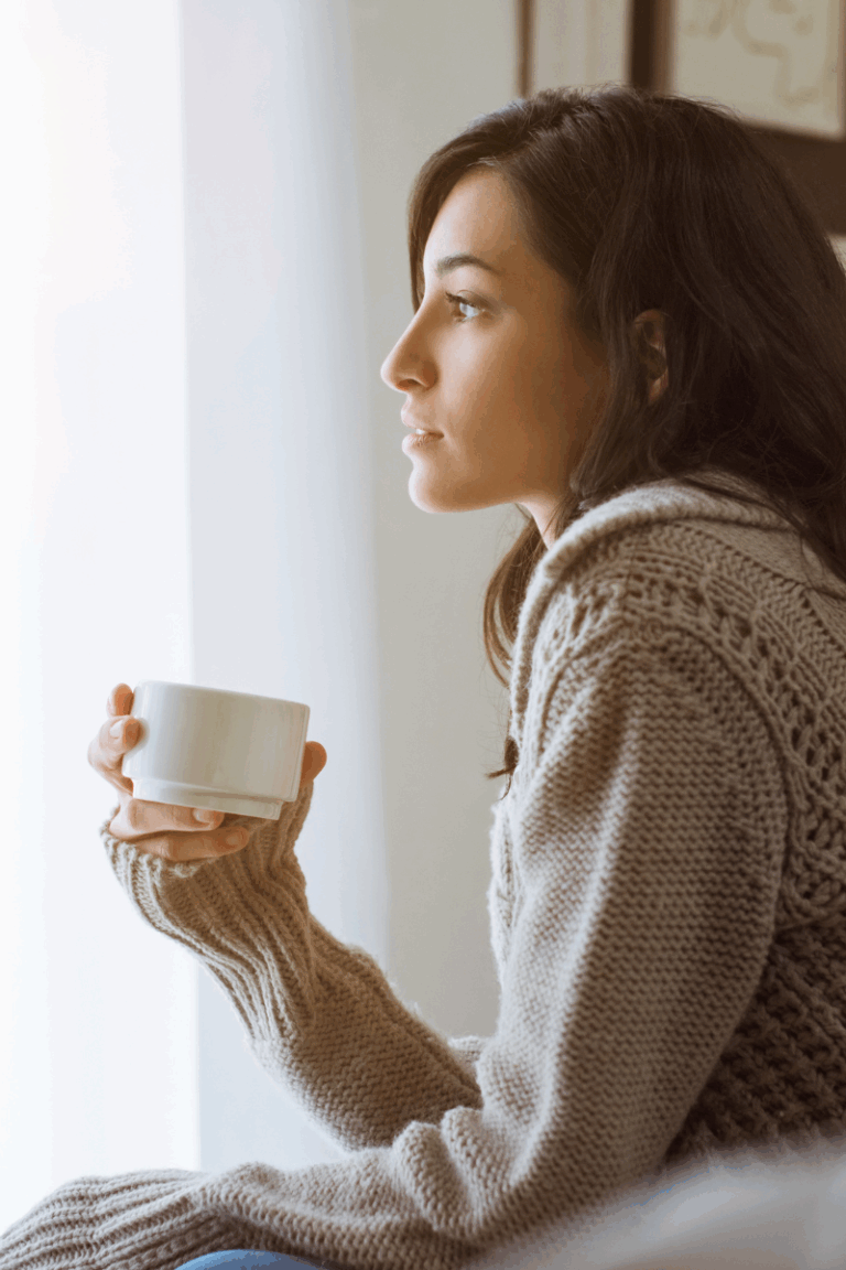 Woman in a cozy sweater holding a coffee mug while gazing out the window, reflecting quietly—capturing the mindset of a procrastinating perfectionist stuck between overthinking and the desire to take action.