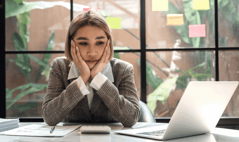 Woman looking overwhelmed at laptop with hands on face, symbolizing uncertainty about how to ask for a testimonial