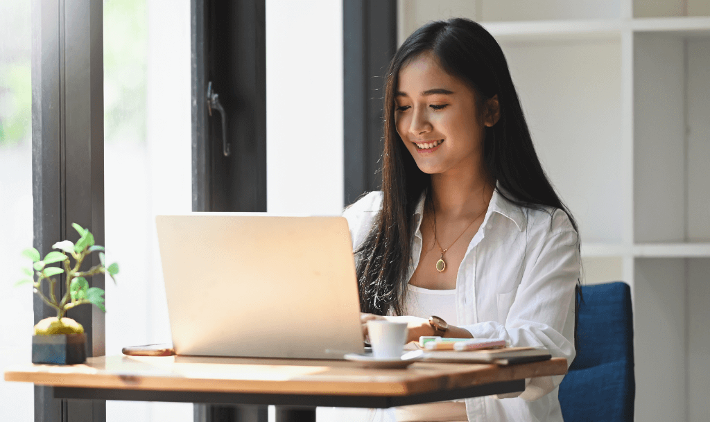 Woman smiling while working on her laptop, reviewing her client onboarding checklist with a cup of coffee beside her.