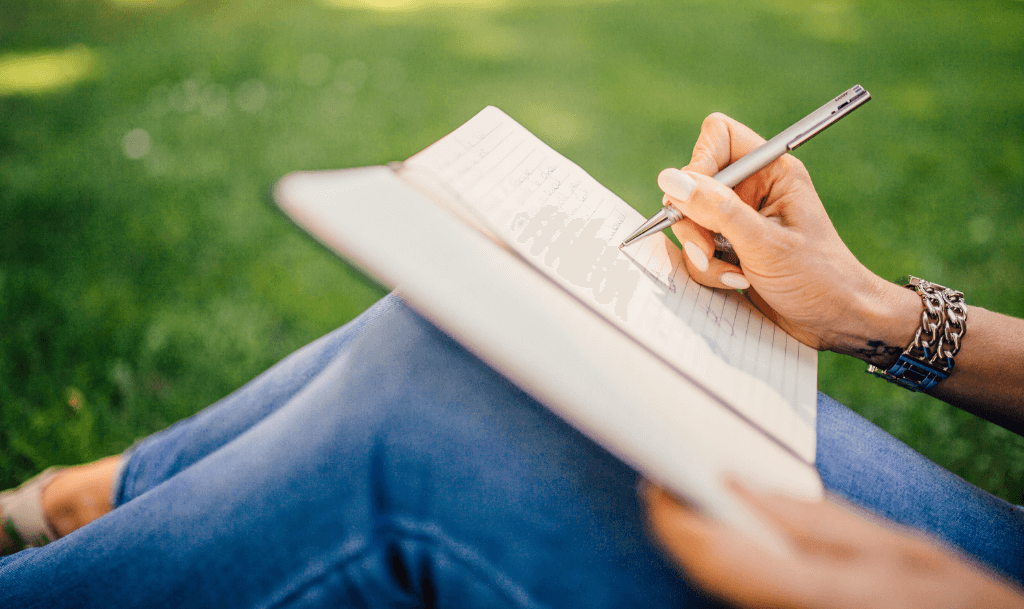 Close-up of a woman writing in a notebook on the grass, peacefully engaging in journaling with prompts. Reflects clarity, mindfulness, and intentional self-reflection in a calm outdoor setting.