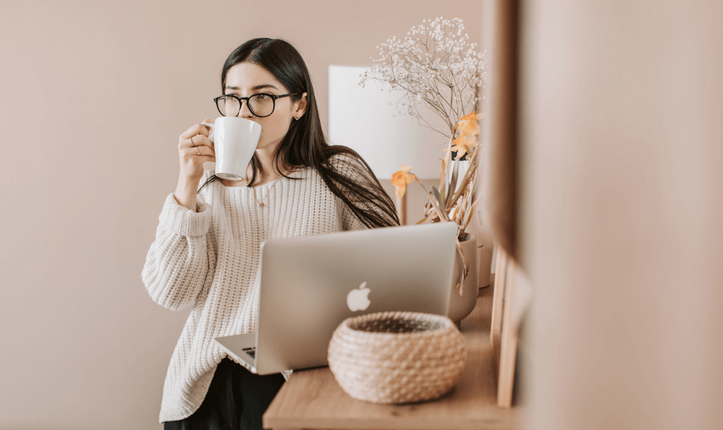 A woman holding a warm mug of coffee during a virtual coffee chat, sitting comfortably at her laptop.